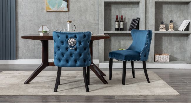 Essgruppe mit rundem Holztisch und zwei getufteten blauen Samtstühlen. Die Stühle sind mit kunstvollen Löwenkopfklopfern und einer Nagelkopfverzierung versehen. Das Zimmer verfügt über eingebaute Regale mit Weinflaschen und Dekor.
