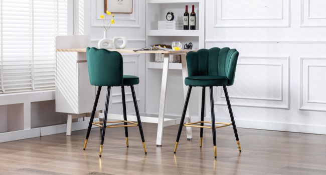 Deux tabourets de bar en velours vert émeraude avec pieds en métal noir et pieds décoratifs dorés, placés sur un comptoir blanc dans un décor intérieur moderne.