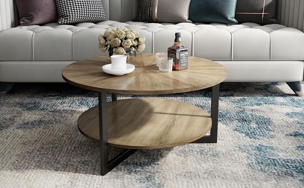 Round wooden coffee table with metal frame and lower shelf. Placed on a blue patterned rug in front of a gray tufted sofa.