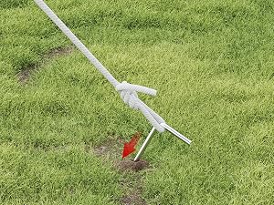 Herramienta o utensilio de jardín de plástico blanco con mango extensible que se muestra en uso en césped verde, con un indicador rojo visible.