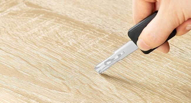 A hand holding a small utility knife with a black handle, cutting into a light wood-grain surface. The blade has multiple notches visible.