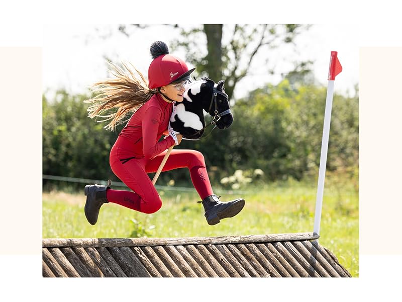 A person jumping over a wooden obstacle while holding a stuffed animal. They wear a red outfit and cap, with a flag visible in the background.