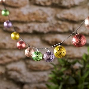 String of colorful, glittery ornament lights hanging against a textured wall. The spherical ornaments come in various colors including red, green, purple, and gold.