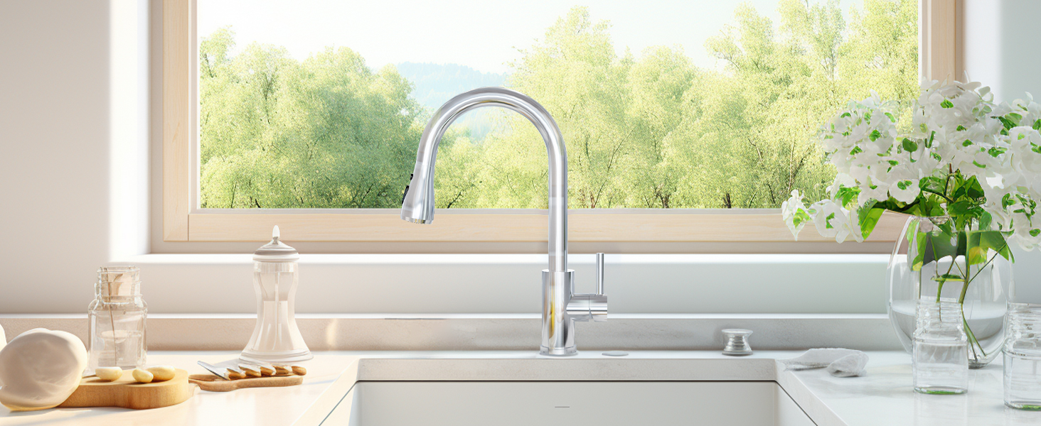 Modern kitchen sink with arched stainless steel faucet, white countertop, and window overlooking greenery. Floral arrangement and kitchen accessories visible.