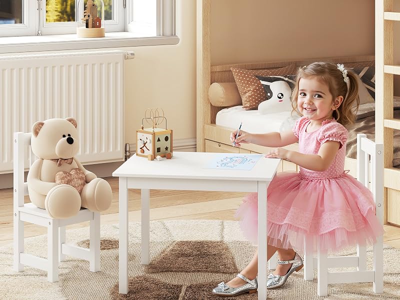 Aménagement de salle de jeux pour enfants avec table et chaises en bois blanc, mis en scène avec des ours en peluche et des jouets. Radiateur blanc et fenêtre visibles en arrière-plan.