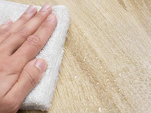 Hand wiping a wooden floor with a white cloth, showing a partial view of fingers and palm against the light-colored wood grain.