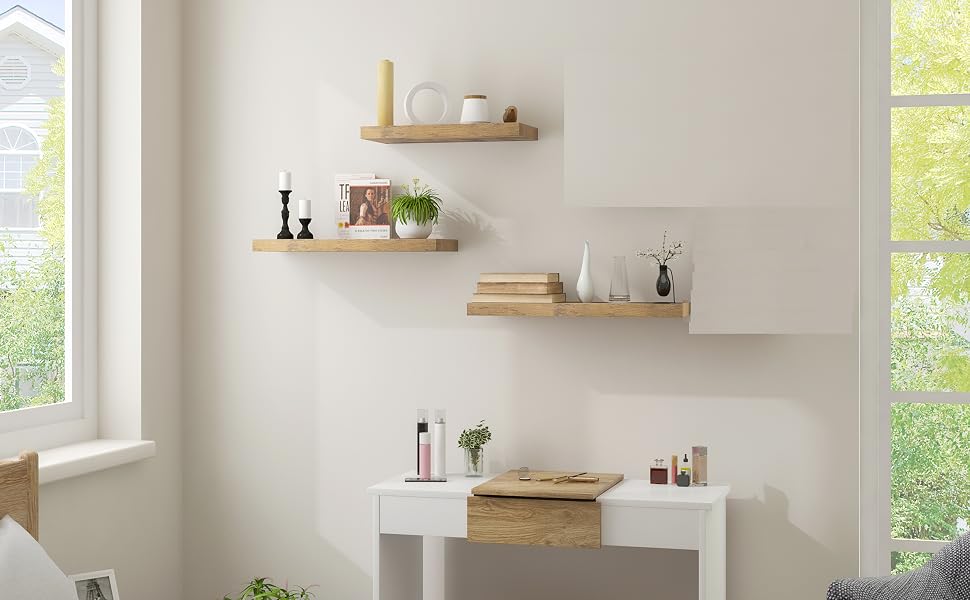 Minimalist bedroom with floating wooden shelves, white vanity table, and stool. Natural light from windows, plants and decor items on shelves and table.