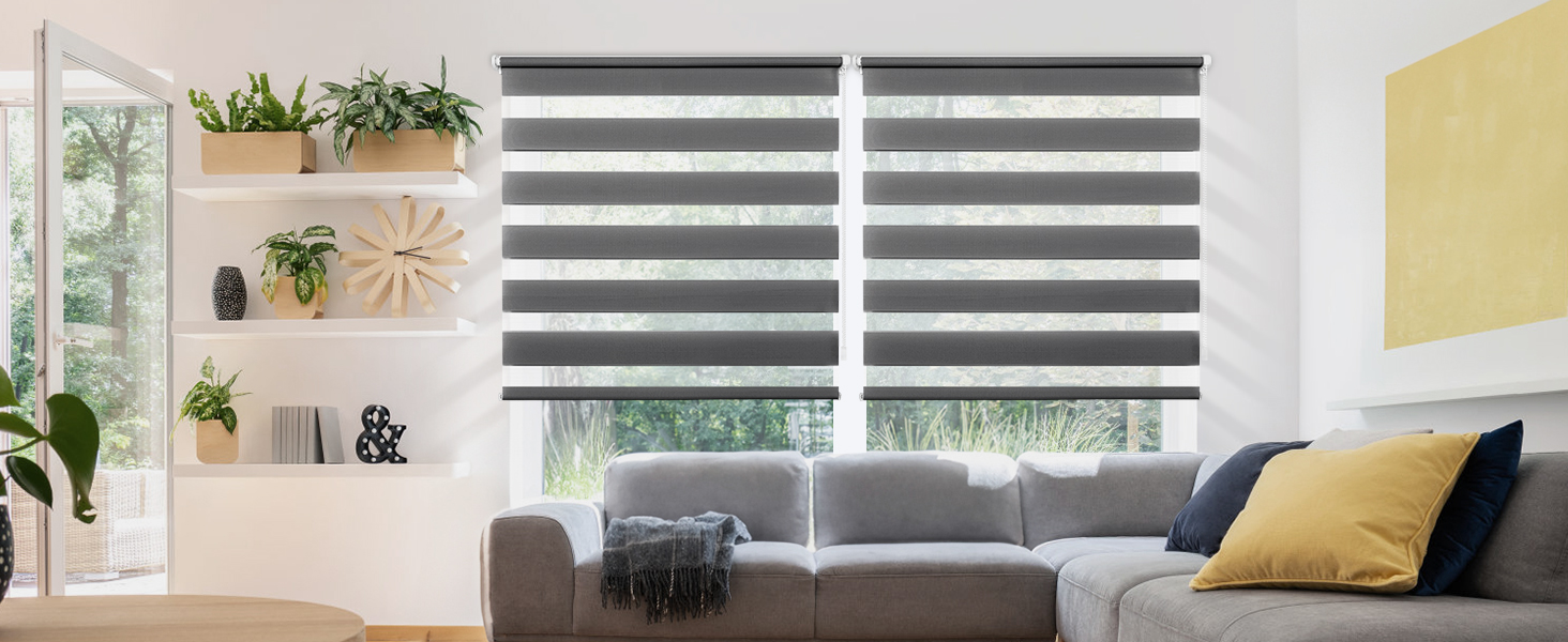 Salon avec canapé sectionnel gris, grandes fenêtres recouvertes de stores enrouleurs rayés gris et blancs. Étagère avec plantes et décoration sur la paroi latérale