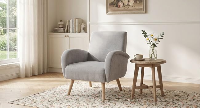 Fauteuil incurvé gris avec pieds en bois positionné à côté d'une petite table d'appoint ronde en bois contenant des fleurs blanches, dans un salon minimaliste lumineux avec une grande fenêtre.