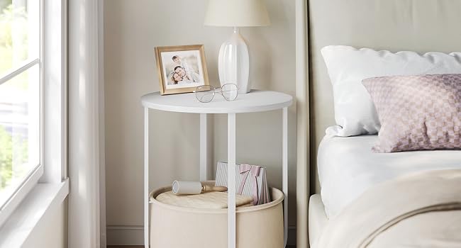 Esquina del dormitorio con mesa auxiliar redonda blanca con lámpara junto a la ventana, decorada con suaves tejidos y almohadas.