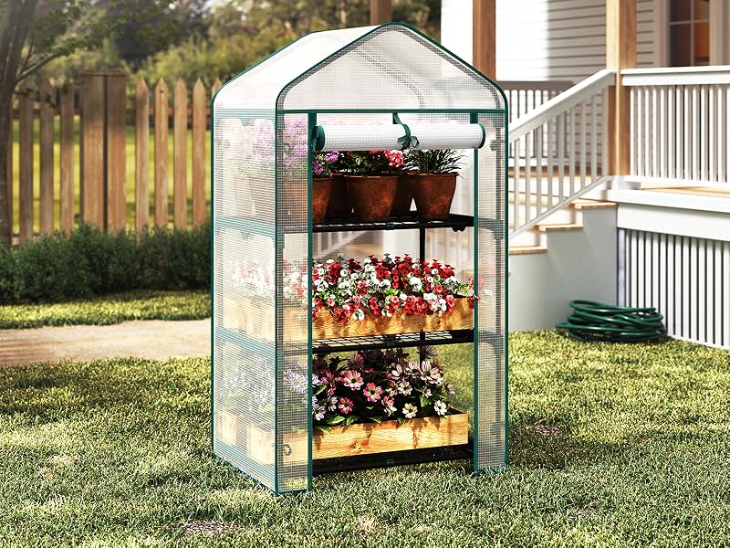 Serra portatile con copertura in plastica trasparente e ripiani multipli, con fiori e piante in vaso, posta sul prato con la casa visibile sullo sfondo.