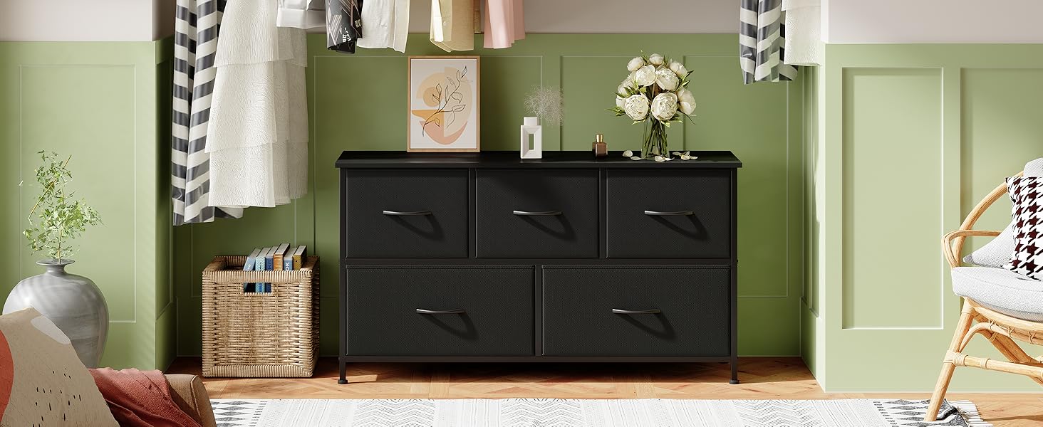 Commode en bois noire à cinq tiroirs dans une pièce aux murs verts. Les objets décoratifs sur le dessus comprennent des fleurs et une photo encadrée. Panier en osier et rideaux visibles à proximité