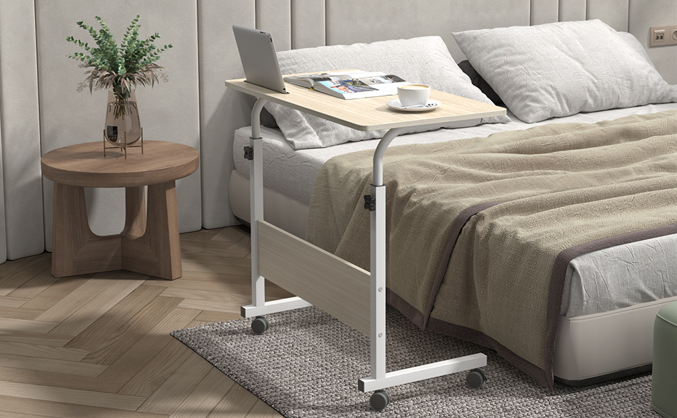 Adjustable over-bed table with white frame and wood-tone top, positioned next to a bed with neutral bedding. Side table and plant visible.