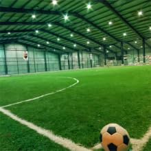 Hallenfußballfeld mit grünem Kunstrasen, weißen Markierungen, LED-Beleuchtung und einem Fußball im Vordergrund. Metall- und Gewebestruktur sichtbar