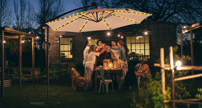 Outdoor evening gathering under illuminated patio umbrella with string lights, showing people socializing around a table.