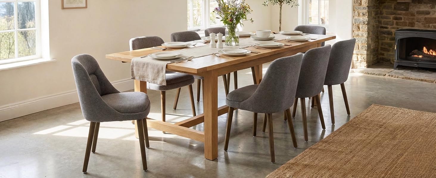 Modern dining room featuring wooden table with cross-beam base, eight grey upholstered chairs, jute area rug, and stone fireplace. Natural light streams through window.