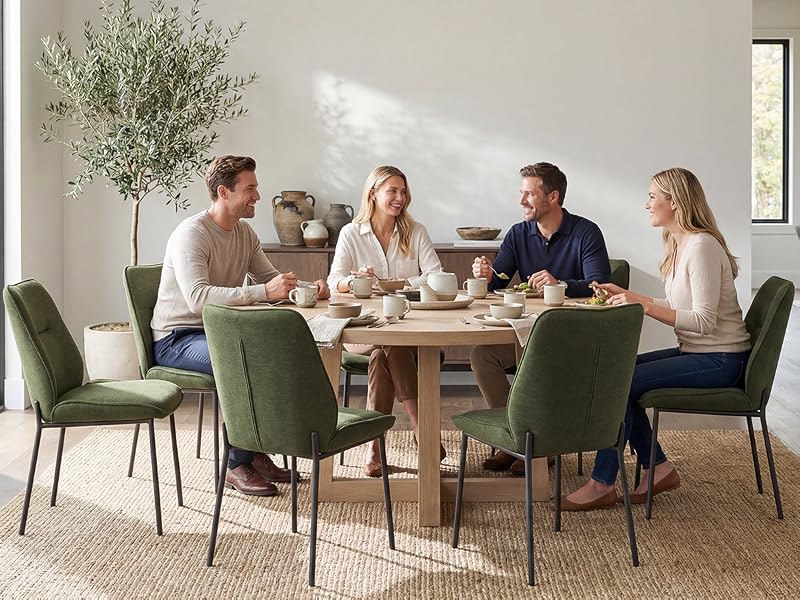 Four people seated around wooden dining table in bright room with green chairs, white flowers in vase, natural daylight from windows, casual dining setting.