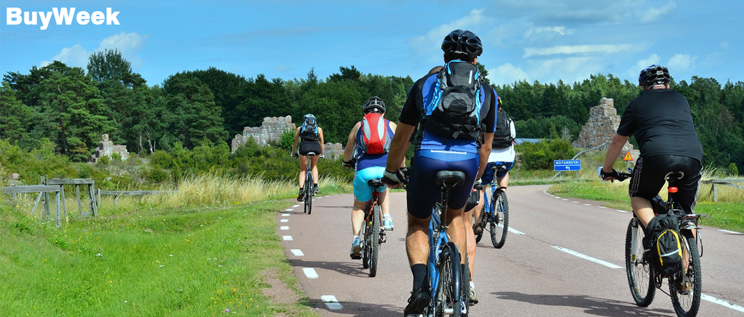 Group of cyclists riding on a paved path through a grassy area. Riders wearing helmets and carrying backpacks.