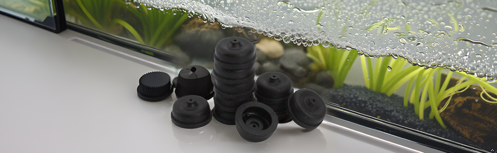 Multiple small black rubber feet or stoppers of various sizes arranged on a reflective surface near a window ledge with plants visible outside.