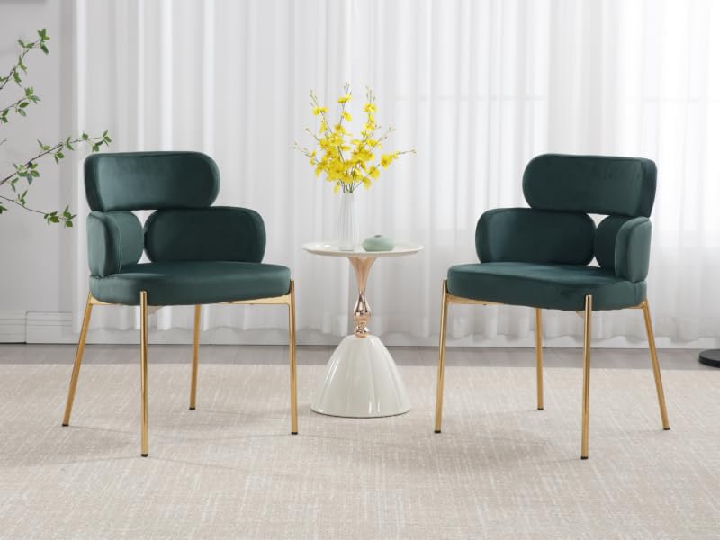 Due sedie imbottite verde scuro con schienali curvi e gambe in metallo color oro. Piccolo tavolino bianco con composizione floreale gialla
