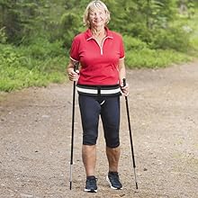 Woman in red top and black pants using walking poles on a dirt path in a forested area.