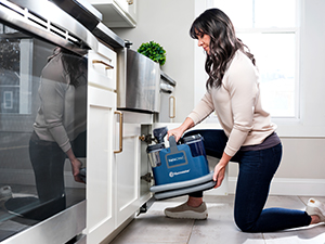 Persona che usa un aspirapolvere portatile blu intenso per pulire l'interno di un mobile da cucina mentre è in ginocchio