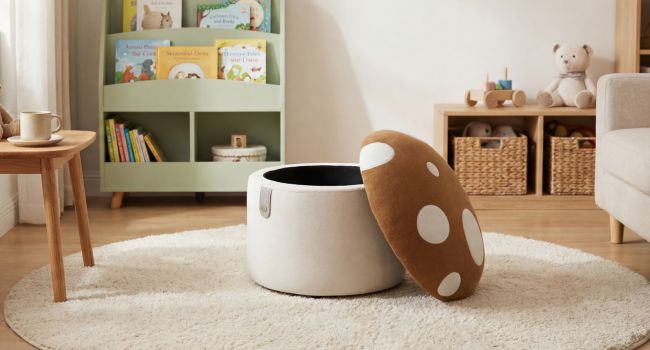 Cream-colored cylindrical fabric storage basket with brown leather handles positioned on light carpet in cozy living room with wooden furniture and green shelving.