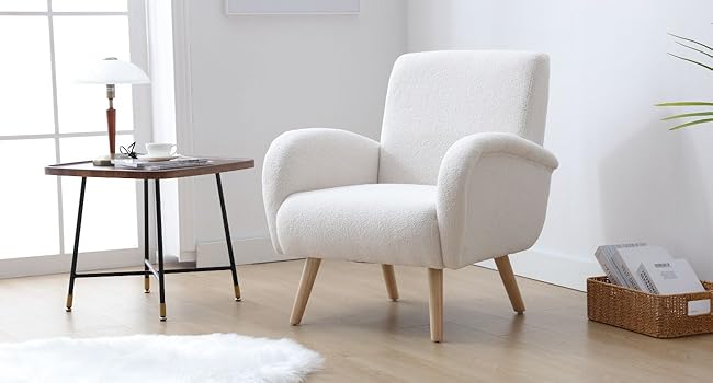 White fabric armchair with curved arms and wooden legs, positioned next to a small round side table in a bright, minimalist living room setting.