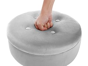 Light gray circular ottoman or footstool with tufted top. A hand is pressing down to demonstrate its soft, cushioned surface.