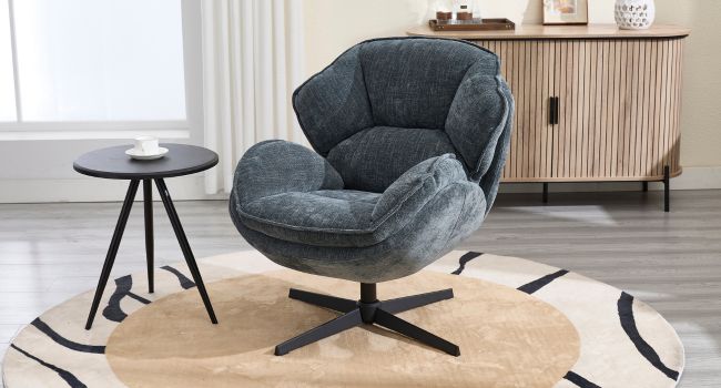 Modern swivel armchair with plush dark grey upholstery, wide curved back, and metal base. Positioned on a round rug next to a small side table in a living room setting.