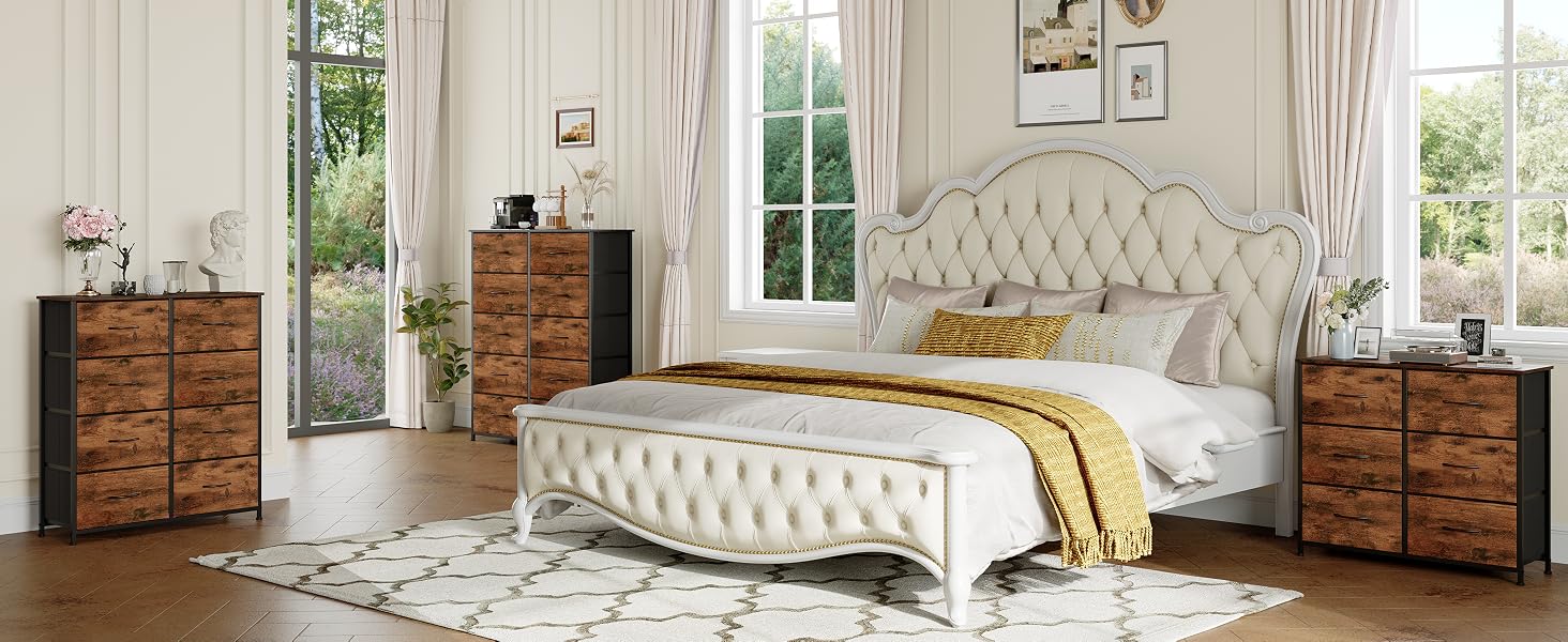 comprenant un lit capitonné blanc rembourré avec tête de lit incurvée, associé à des commodes rustiques en bois. Cette chambre est dotée de murs blancs, de grandes fenêtres et d'un tapis à motifs