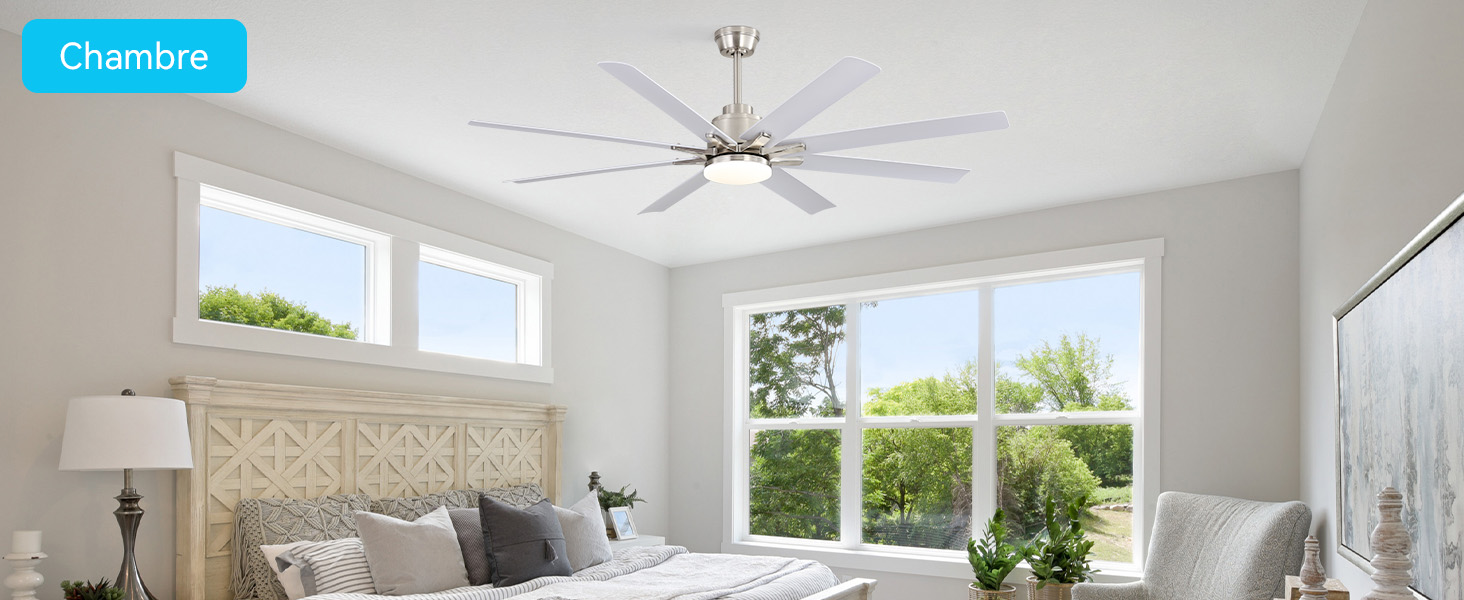 Le texte indique « Chambre ». Intérieur lumineux de la chambre avec ventilateur de plafond, grandes fenêtres, lit avec tête de lit décorative, lampes de chevet, plantes en pot et murs blancs.