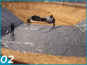 Sitio de construcción con un gran pozo excavado. Trabajadores colocando láminas de plástico gris sobre suelo arenoso, cubriendo parte del área de excavación.