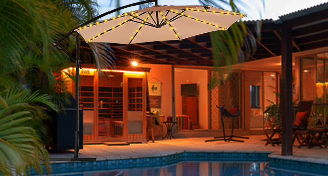 Large beige patio umbrella with LED string lights illuminated at night, positioned beside swimming pool area with tropical landscaping.