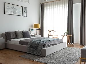Camera da letto moderna con letto imbottito grigio, tavolino in legno, lampada gialla e ampie finestre con tende grigie. Opere d'arte incorniciate su pareti bianche