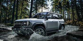 Robusto SUV che guida in acque poco profonde in una foresta. Esterno grigio argento con design squadrato. Pneumatici di grandi dimensioni e altezza da terra elevata per prestazioni fuoristrada.