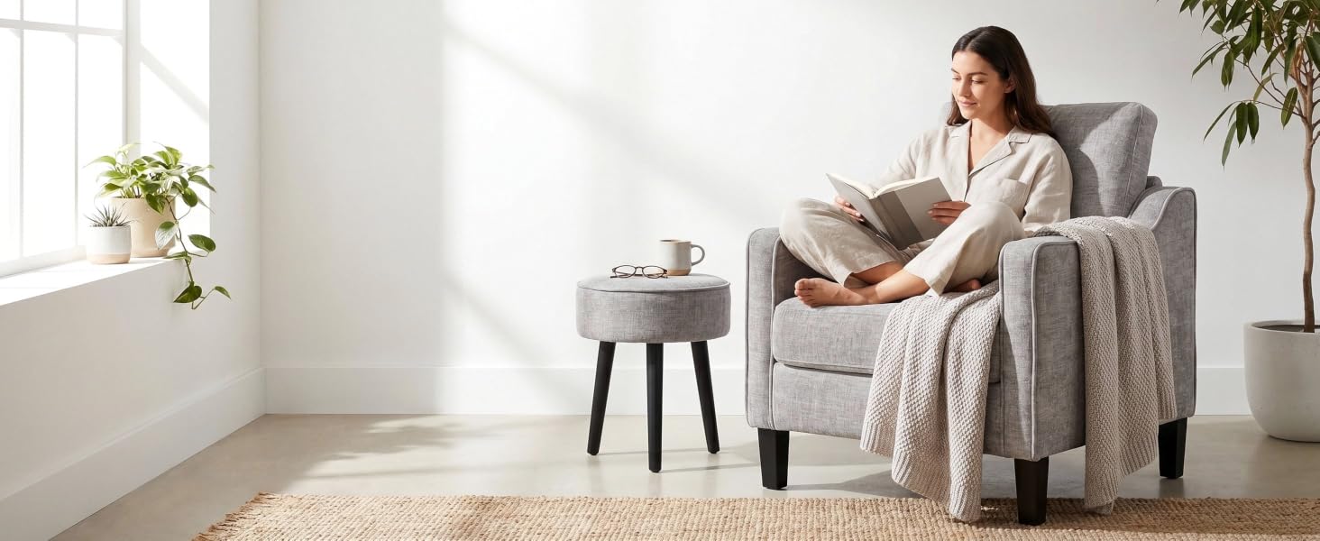 Woman seated in beige armchair with laptop in minimalist living room featuring white walls, small side table, potted plants, and wooden flooring.