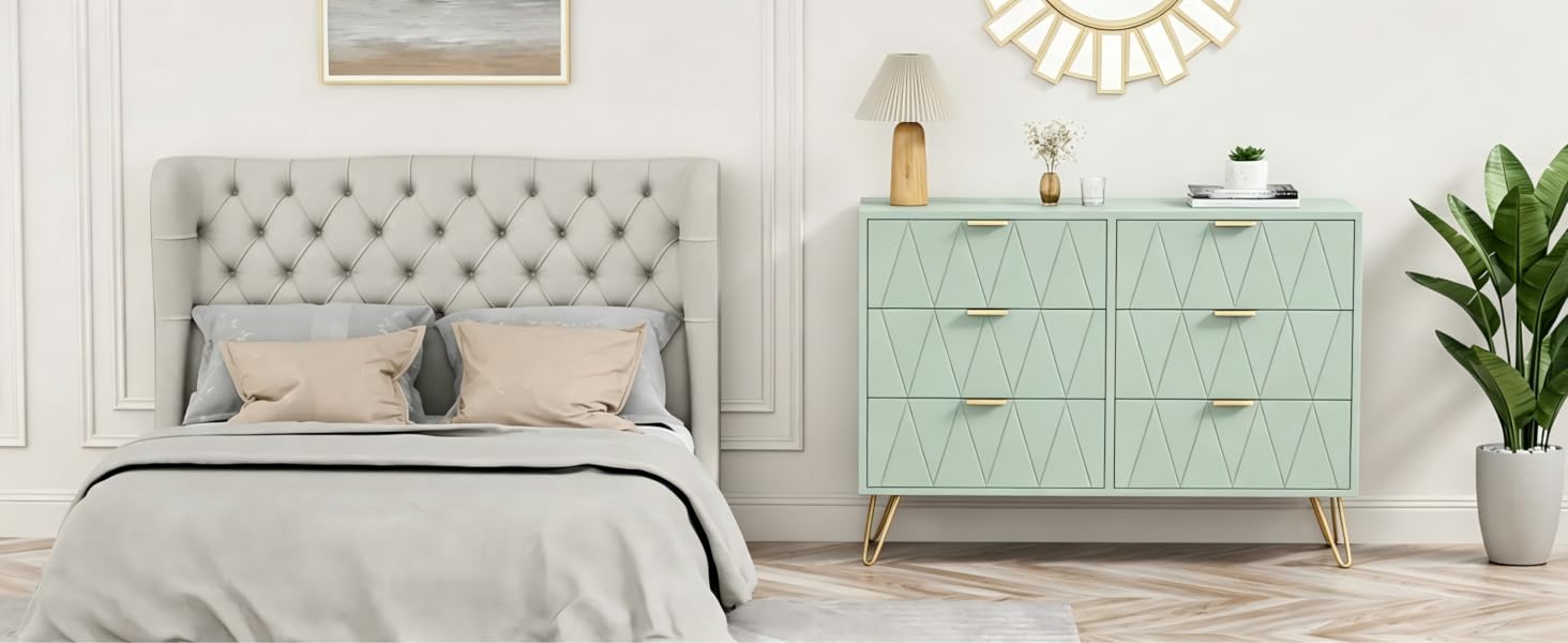 L'intérieur de la chambre comprend un lit rembourré avec tête de lit grise, une commode verte avec poignées de tiroir, des plantes en pot et un agencement de meubles de chambre dans une palette de couleurs douces et neutres.