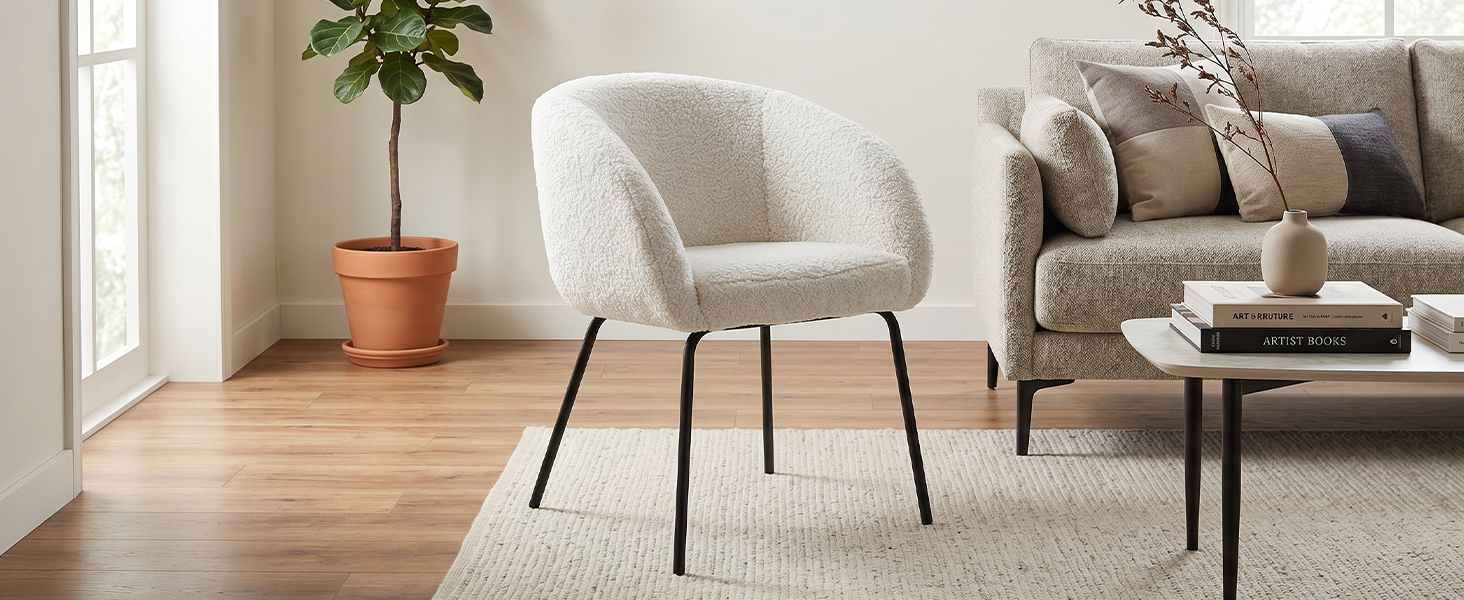 Modern living room interior featuring light gray upholstered armchair with black metal legs, beige sofa, wooden flooring, potted plant, and natural window lighting.