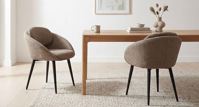 Deux chaises longues rembourrées brunes avec pieds en métal noir positionnées sur un bureau en bois dans un bureau à domicile neutre ou un espace de vie aux murs blancs et à la décoration minimale.