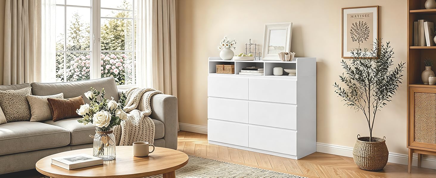 Comò in legno bianco con più cassetti posizionato nel salotto moderno. Arredato con oggetti decorativi tra cui piante in vaso, fiori in vaso e mobili beige neutri visibili sullo sfondo.
