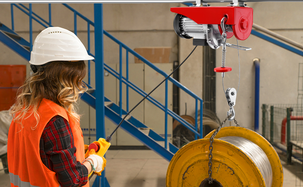Paranco elettrico industriale con custodia rossa che solleva una grande bobina o tamburo giallo. Operaio con giubbotto di sicurezza arancione e elmetto bianco che opera