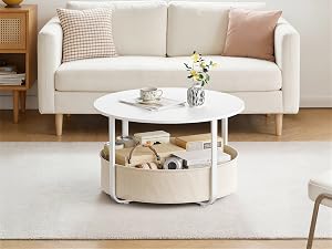 Cremefarbenes Polstersofa mit gepolsterten Armlehnen im hellen Wohnzimmer mit weißem rundem Couchtisch darunter, geflochtenem Aufbewahrungskorb darunter, Fenster mit Tageslicht dahinter, minimalistischer Wohnstil.