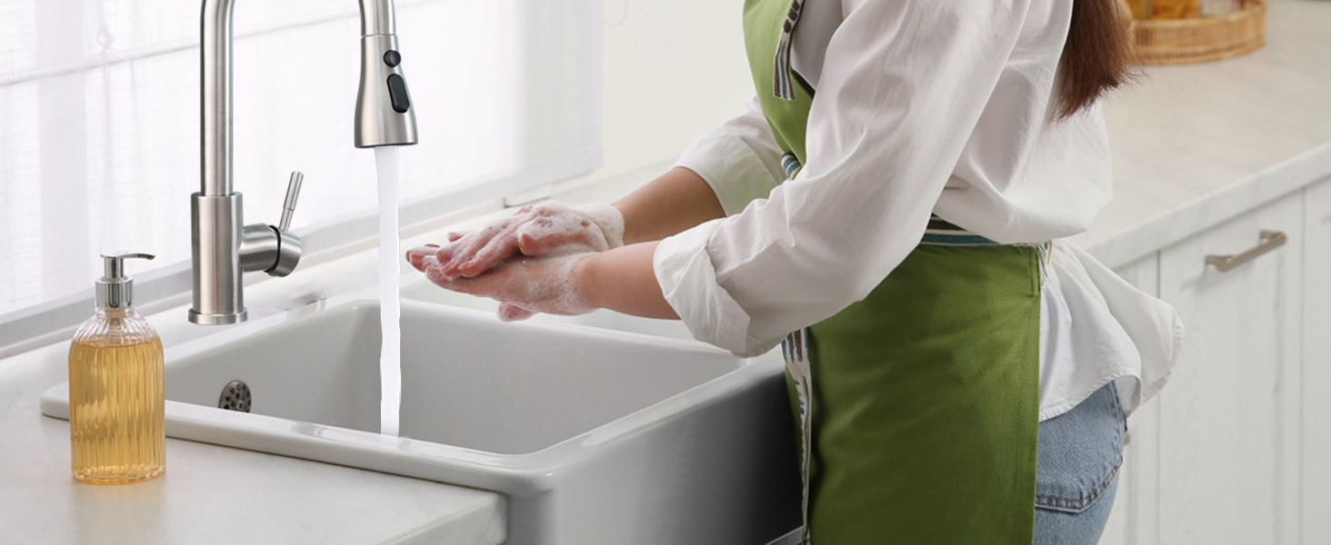 Person wearing white top and green apron washing hands in a stainless steel kitchen sink. A wooden brush is visible near the faucet.