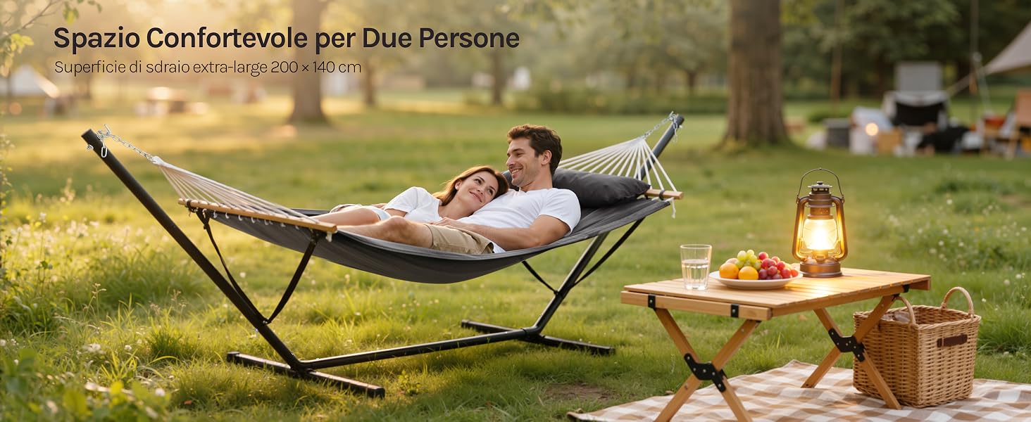 Il testo recita «Spazio Confortevole per Due Persone». Amaca da esterno con lanterna e attrezzatura da picnic su un piccolo tavolo di legno al crepuscolo.