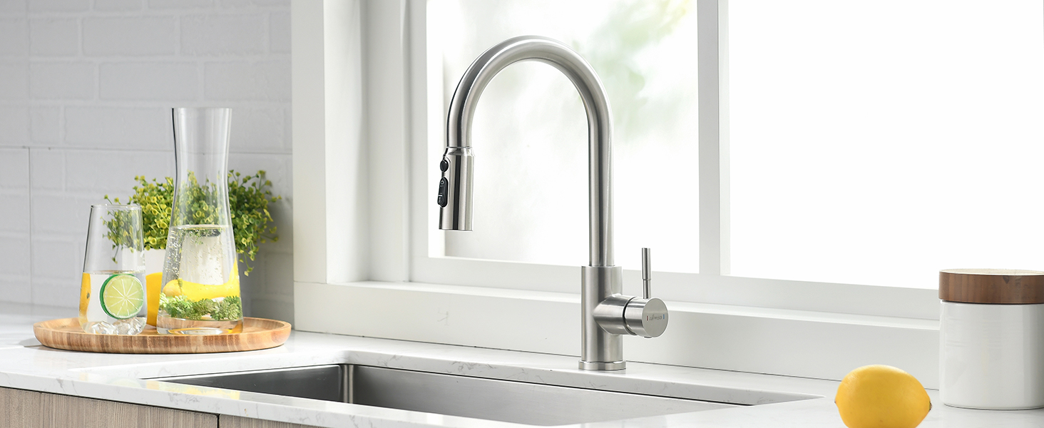Modern stainless steel kitchen faucet with pull-down sprayer installed over white sink, bright window setting with herbs visible.