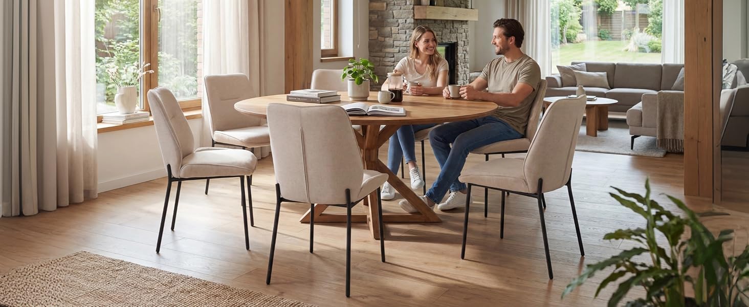 Interno della sala da pranzo dai toni caldi con tavolo in legno con quattro sedie imbottite neutre, finestre illuminate dal sole con tende trasparenti, pareti color crema, piante in vaso verdi e luce naturale che creano uno spazio luminoso e invitante.