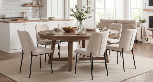 Interno della sala da pranzo con tavolo rotondo in legno con sedie imbottite color crema, mobili da cucina bianchi visibili sullo sfondo, centrotavola per piante in vaso, tappeto, illuminazione naturale brillante dalla finestra.