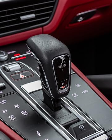 Close-up of modern car gear shifter with electronic display in black finish, set against red interior trim and dashboard with illuminated control buttons.