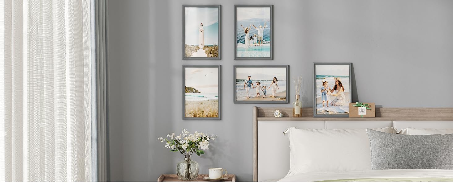 Interno grigio della camera da letto con tende trasparenti bianche, disposizione di opere d'arte incorniciate a parete sopra il comò bianco, oggetti decorativi e vaso sulla superficie.